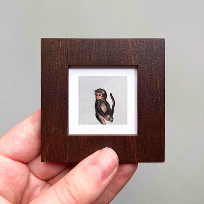 A hand is holding a small wooden frame containing a framed image of a monkey.