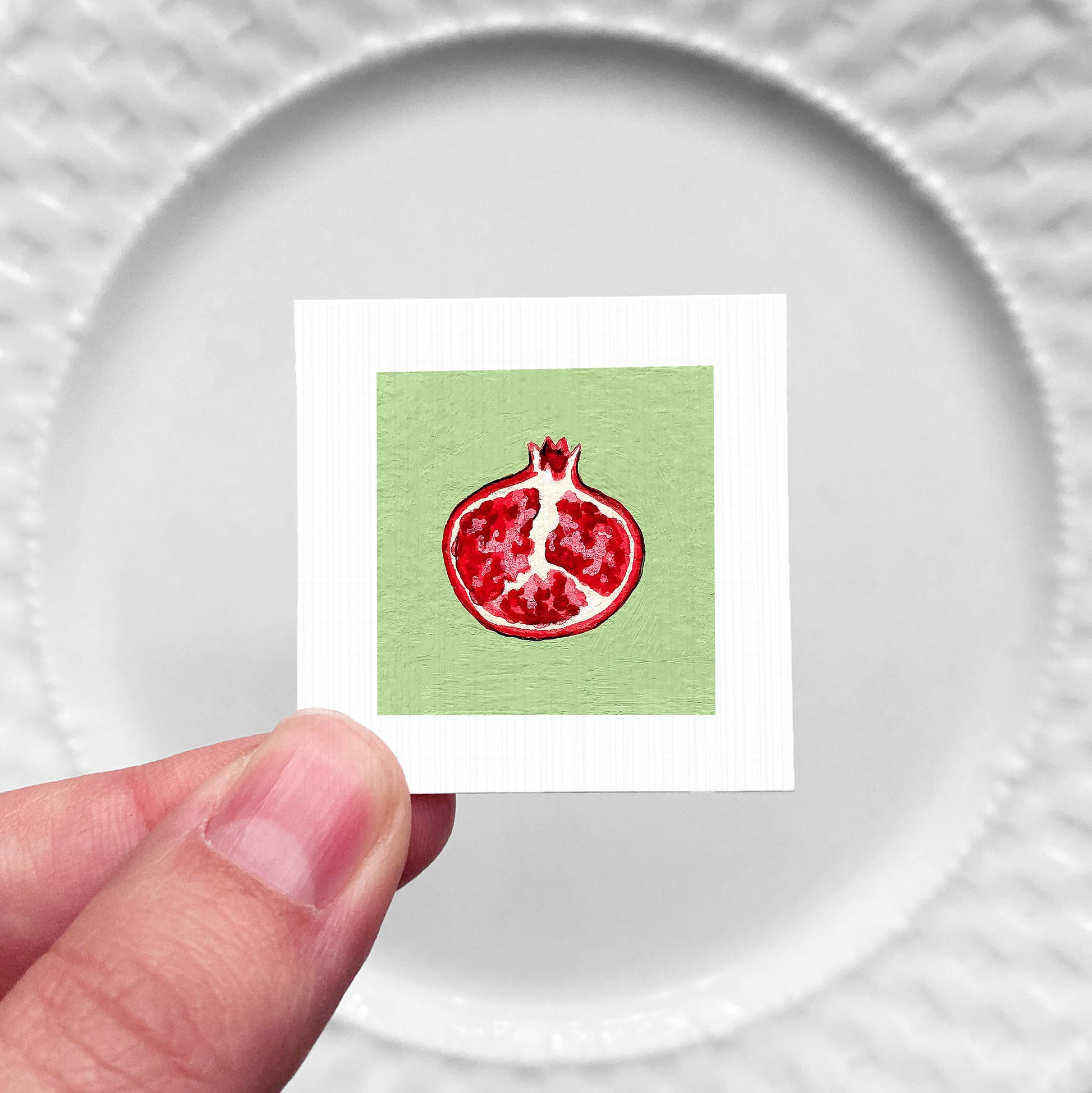 A hand is holding a small square with a pomegranate image on it, against a white background.