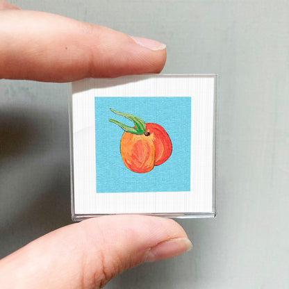 A person's hand holding a small square with a vibrant illustration of a peach on a blue background.
