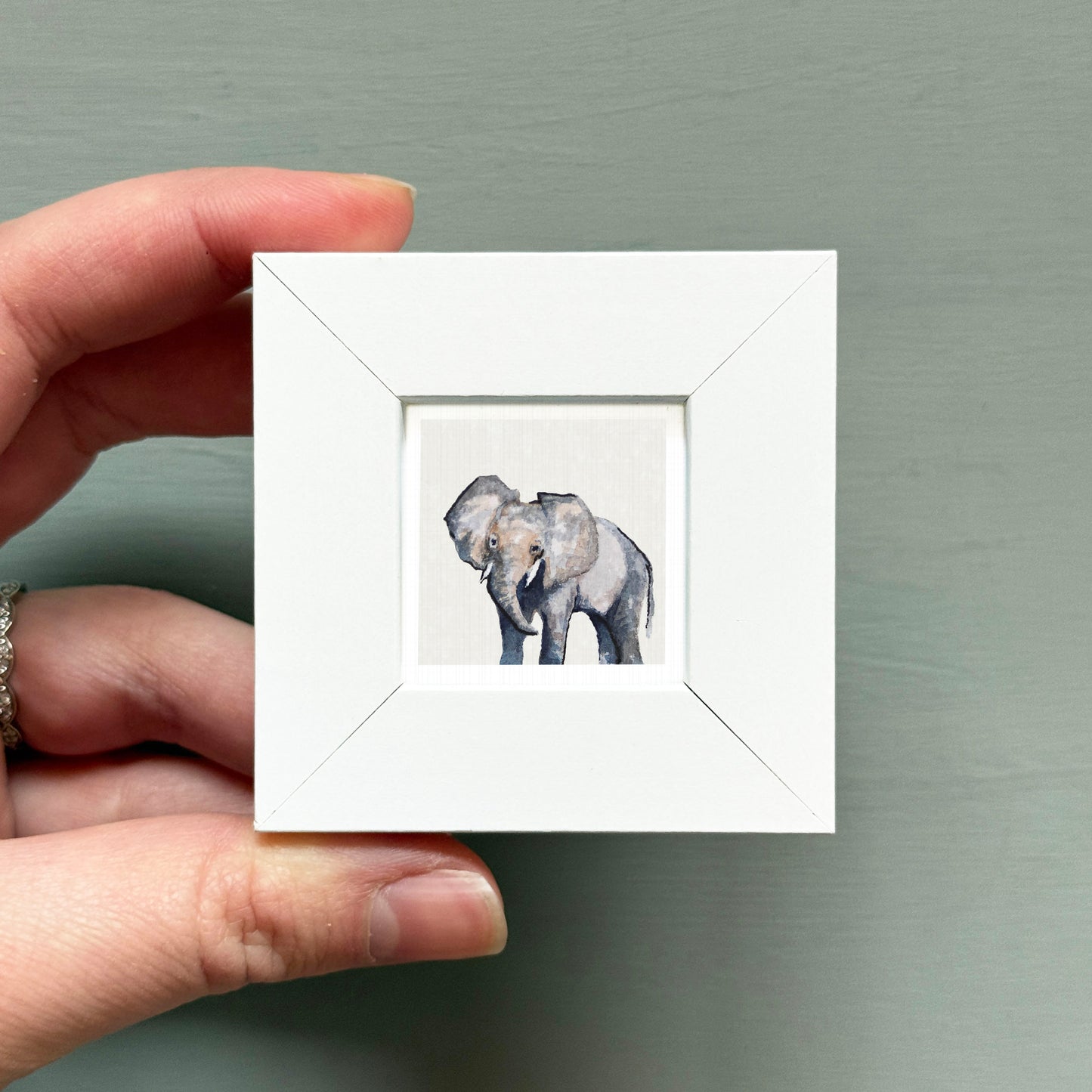 A hand is holding a small white square frame containing a painting of two elephants.