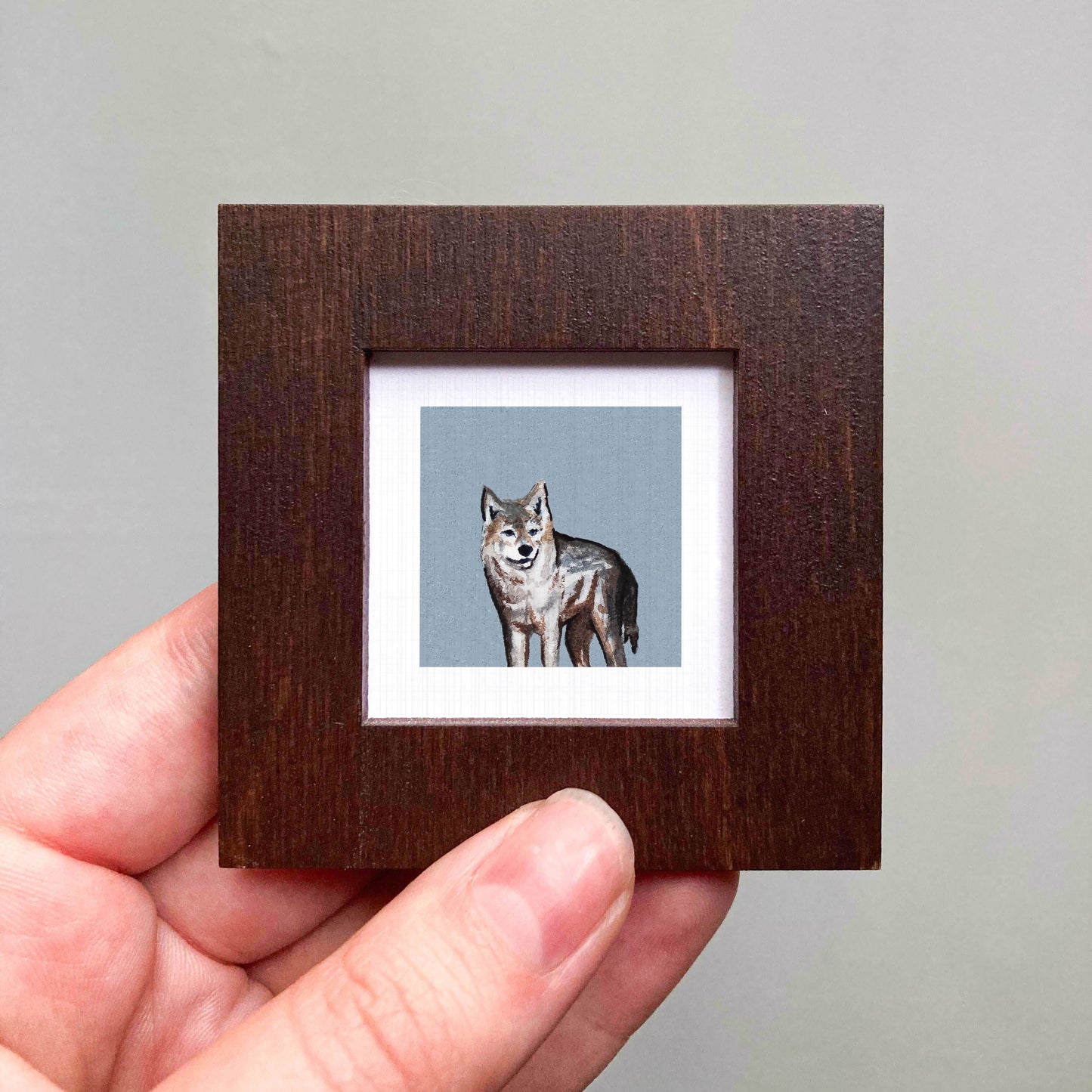 A hand is holding a small wooden frame containing a framed painting of a wolf.