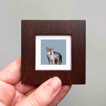 A hand is holding a small wooden frame containing a framed painting of a wolf.