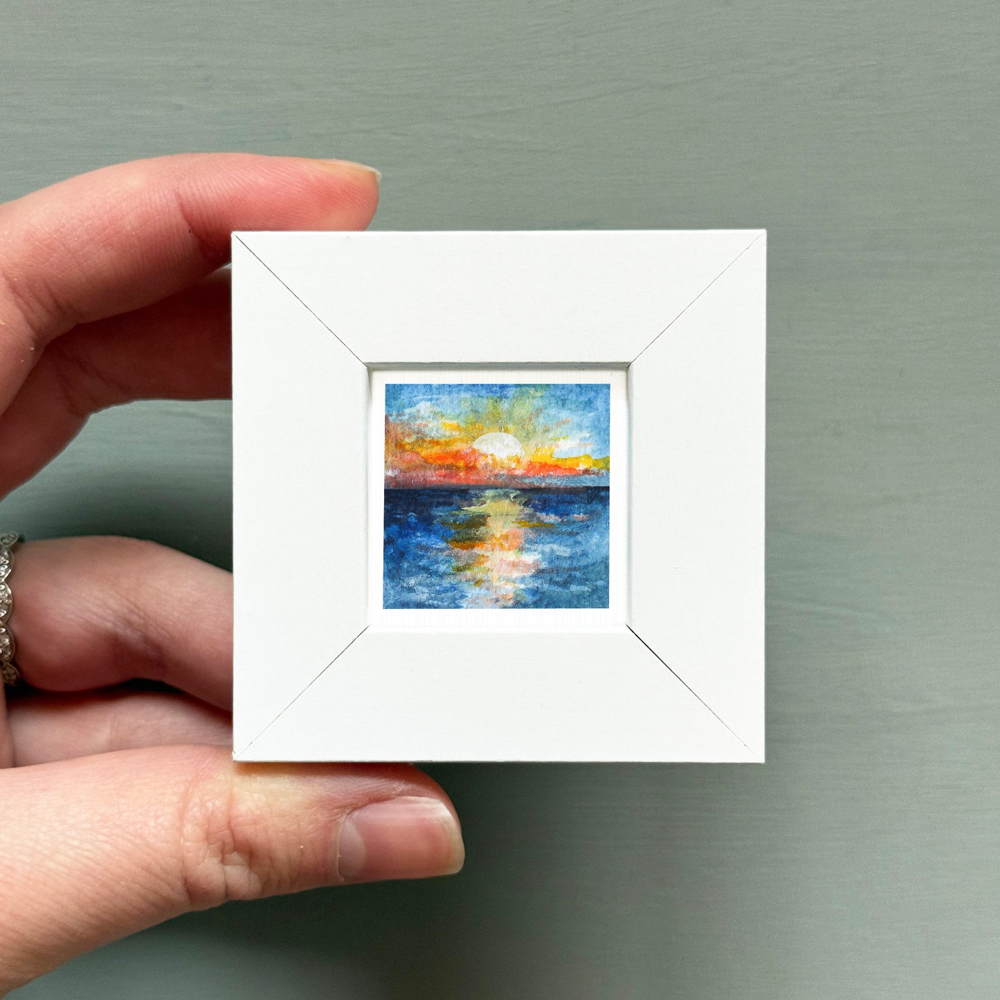 A hand is holding a small white square frame containing a painting of a sunset over a body of water.