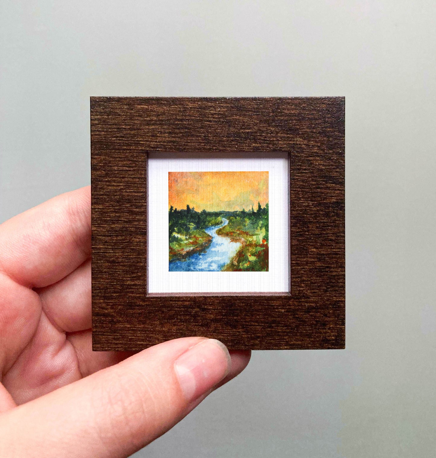 A hand is holding a small, square-shaped wooden frame containing a painting of a river and trees.