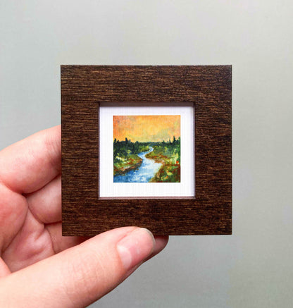 A hand is holding a small, square-shaped wooden frame containing a painting of a river and trees.