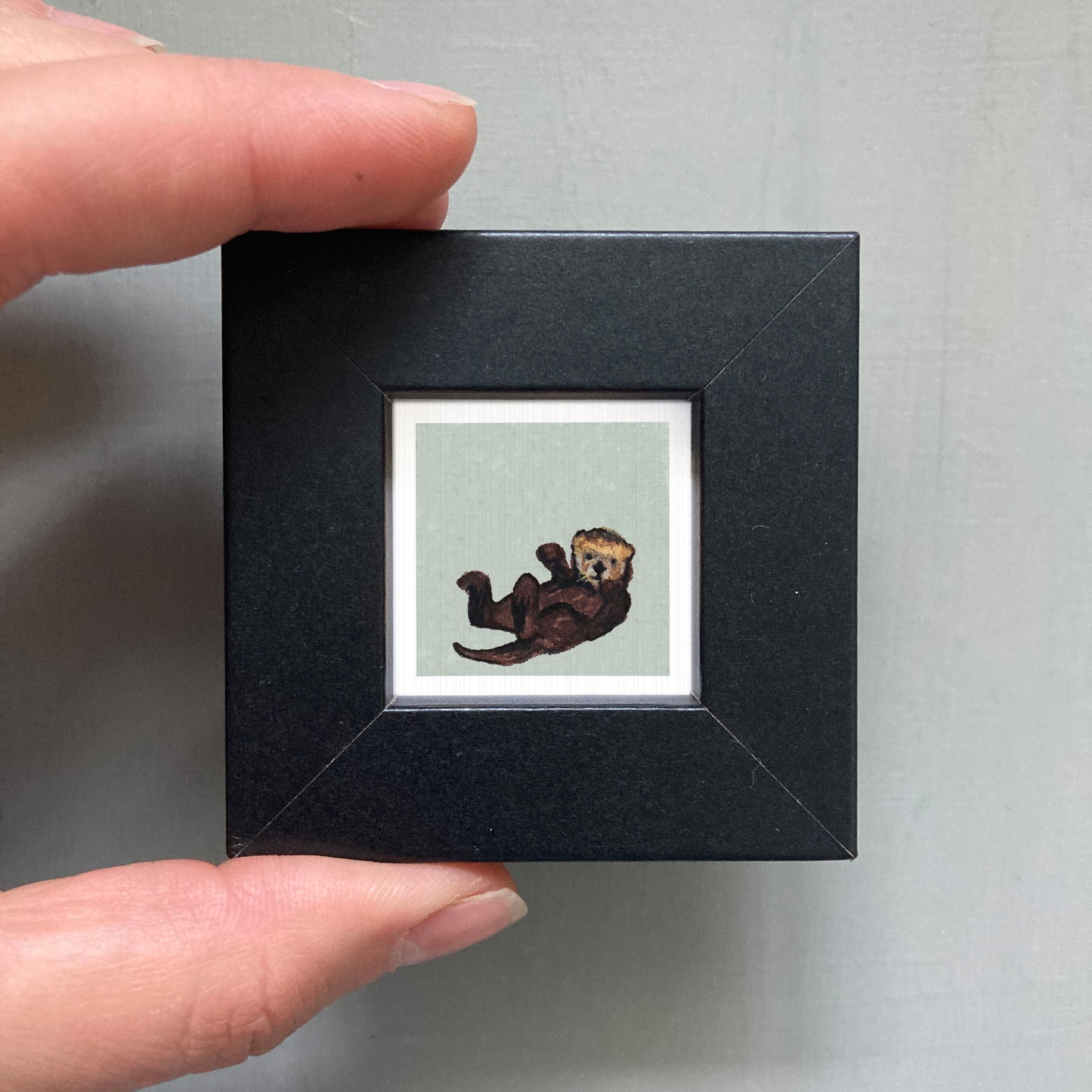 A person's hand is holding a small black frame containing a framed image of an otter.