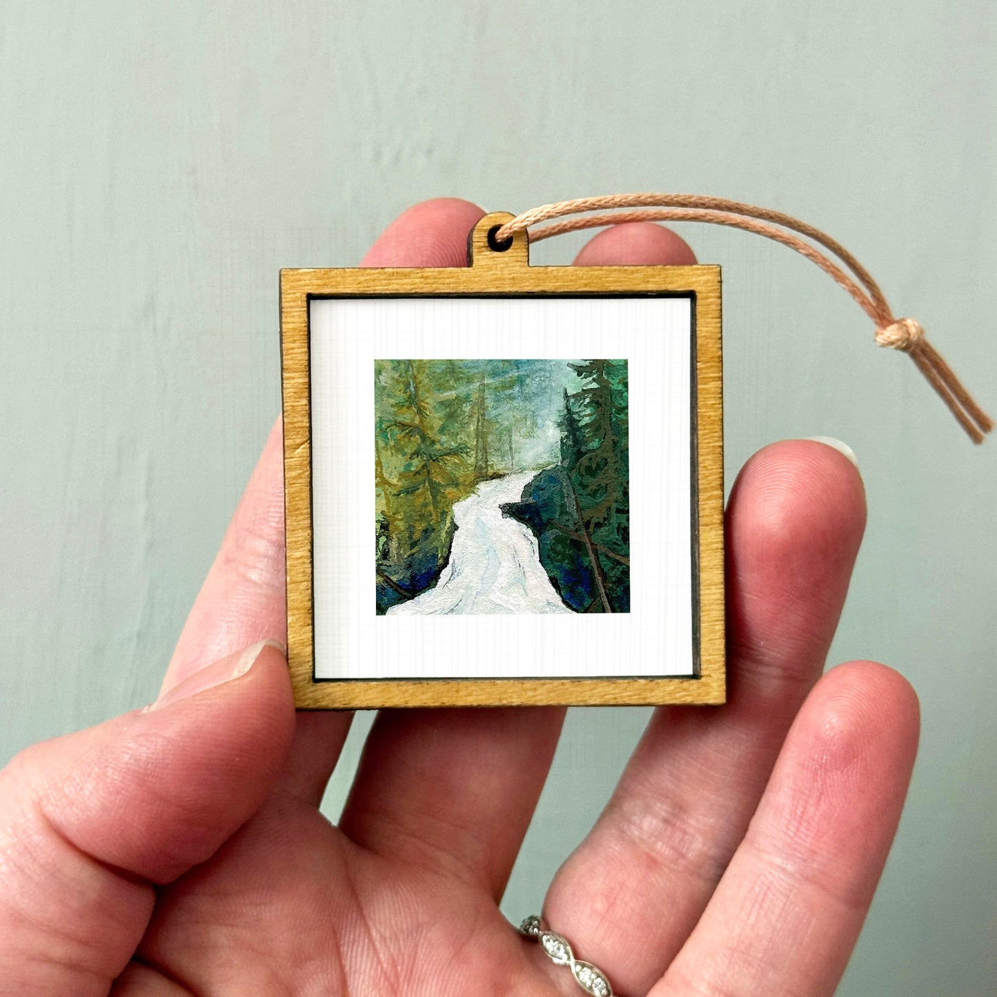 A person's hand holding a small wooden frame containing a painting of a forest scene.