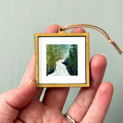 A person's hand holding a small wooden frame containing a painting of a forest scene.