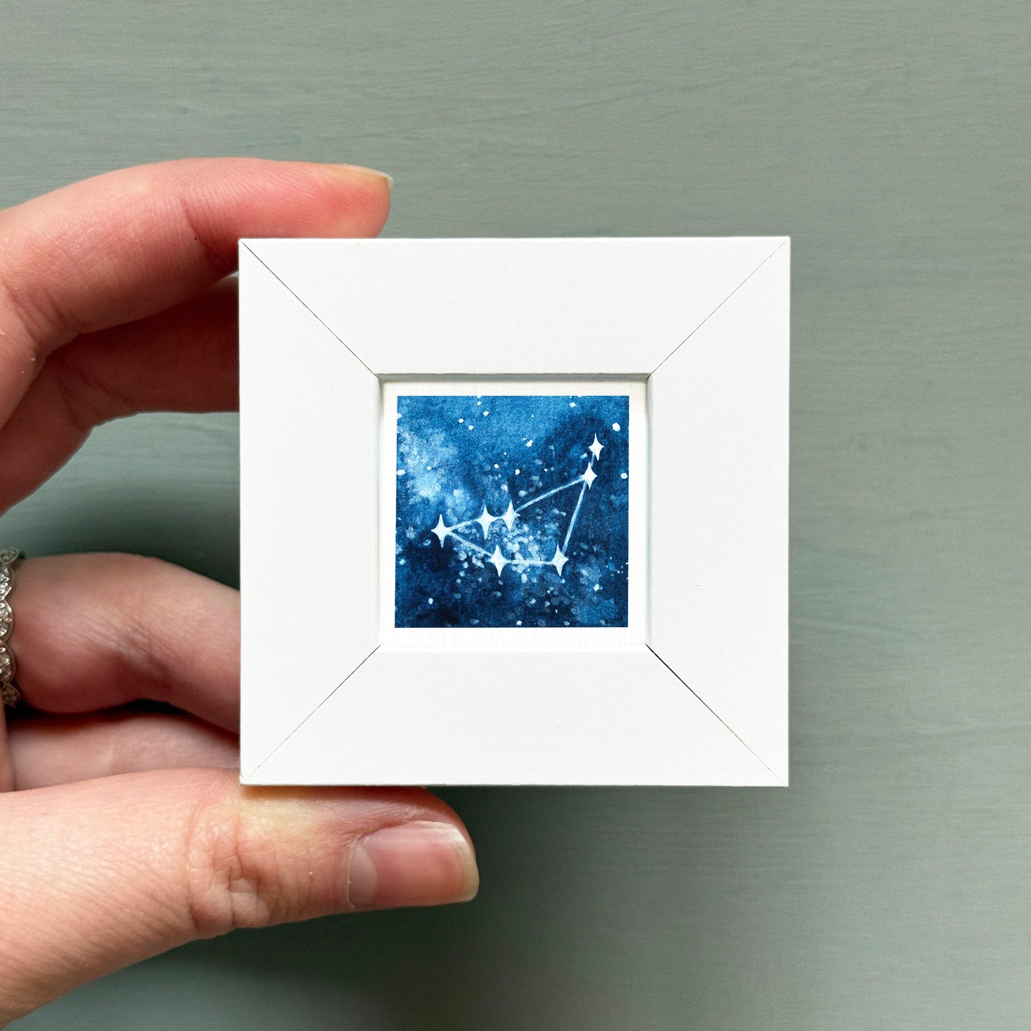 A hand is holding a small white square frame containing a painting of a starry night sky.