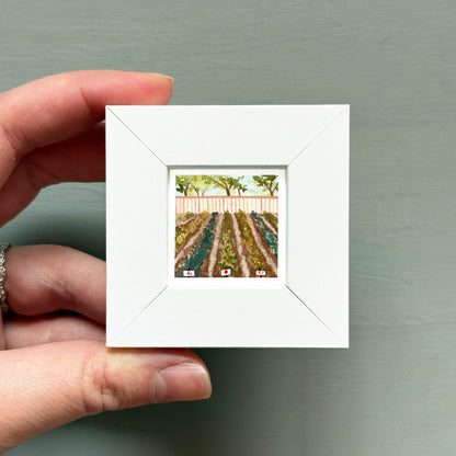 A hand is holding a small white square frame containing a painting of a field with trees and a fence.