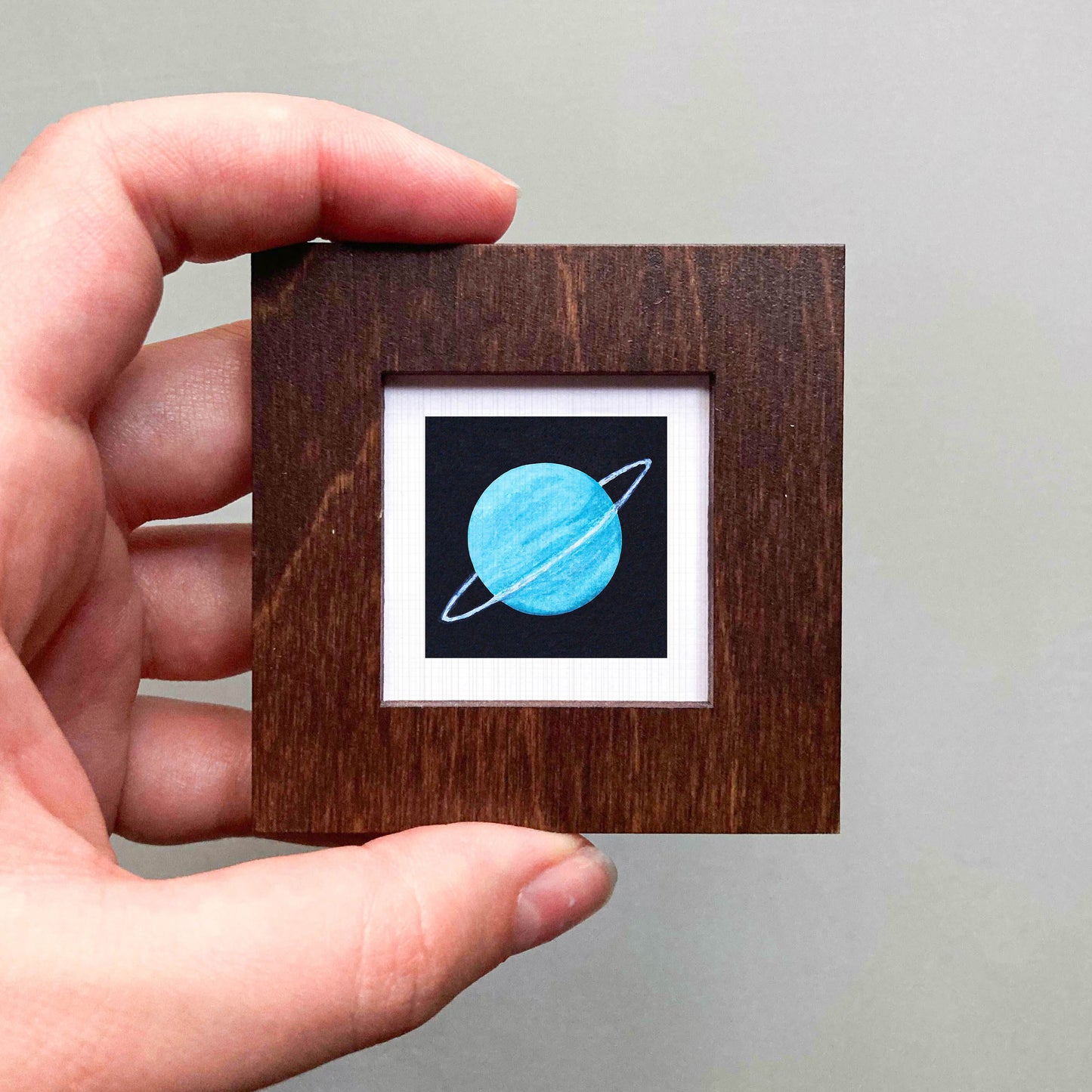 A hand is holding a small wooden frame containing a framed image of a blue planet with a ring around it.