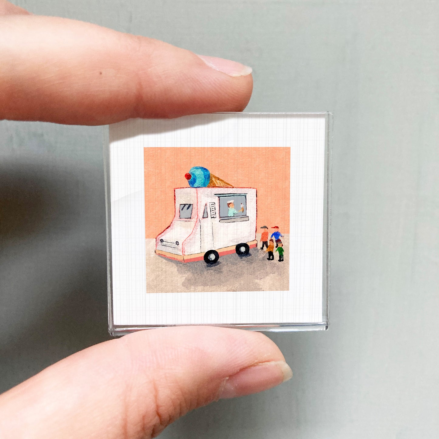 A person's hand is holding a small square picture depicting a colorful ice cream truck with people around it.