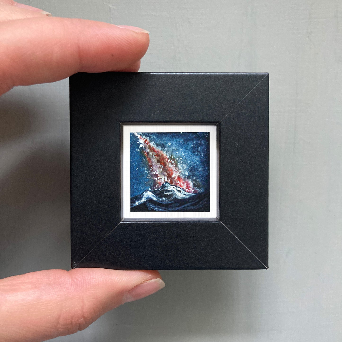 A person's hand is holding a small, square-shaped black frame containing a vibrant, colorful painting of a crashing wave.