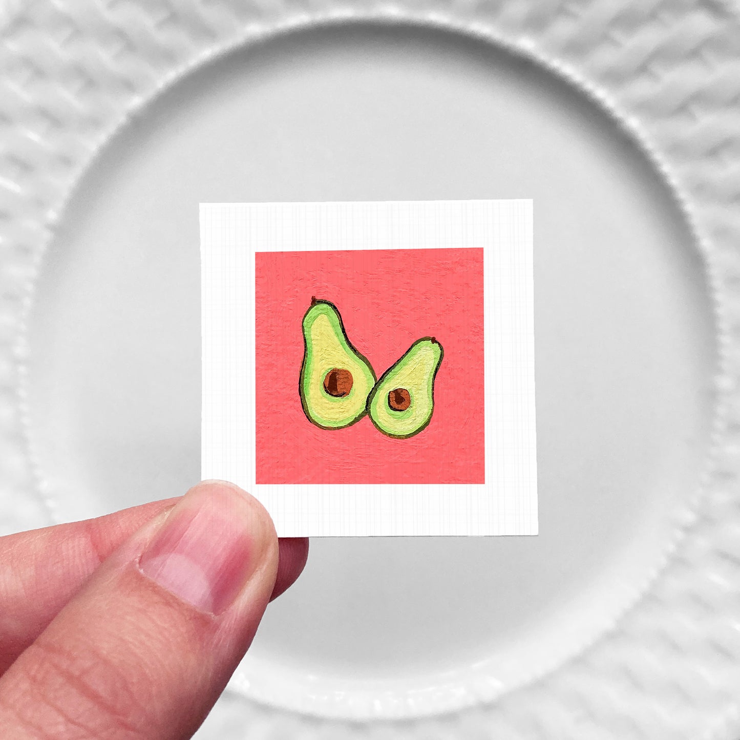 A hand is holding a small square with an illustration of two avocados on a red background.