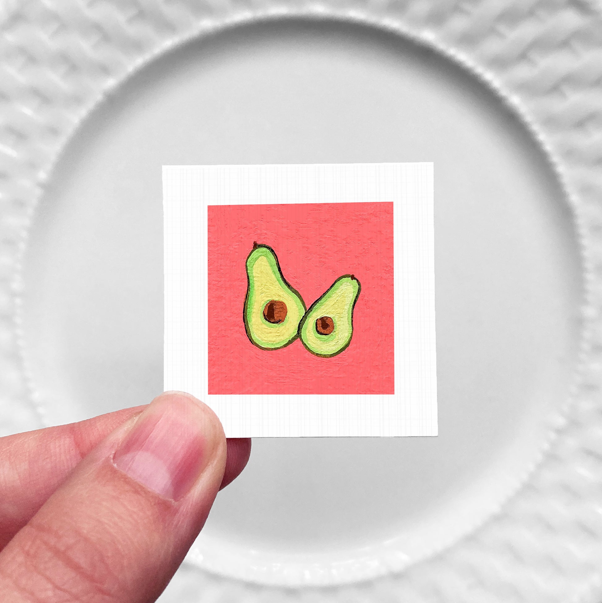 A hand is holding a small square with an illustration of two avocados on a red background.