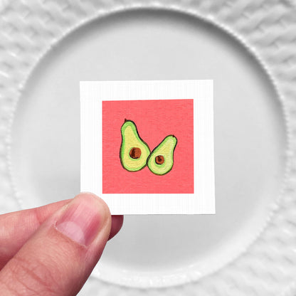 A hand is holding a small square with an illustration of two avocados on a red background.