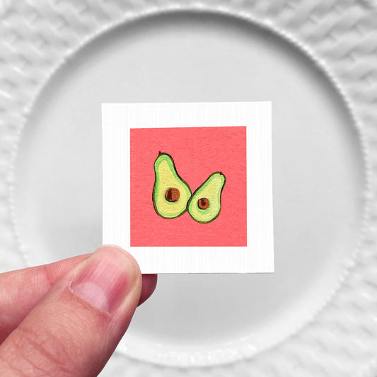 A hand is holding a small square with an illustration of two avocados on a red background.