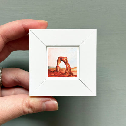 A hand is holding a small white square frame containing a painting of a natural rock formation.