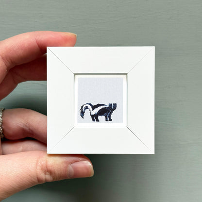 A hand is holding a small white square frame containing an embroidered image of a skunk.