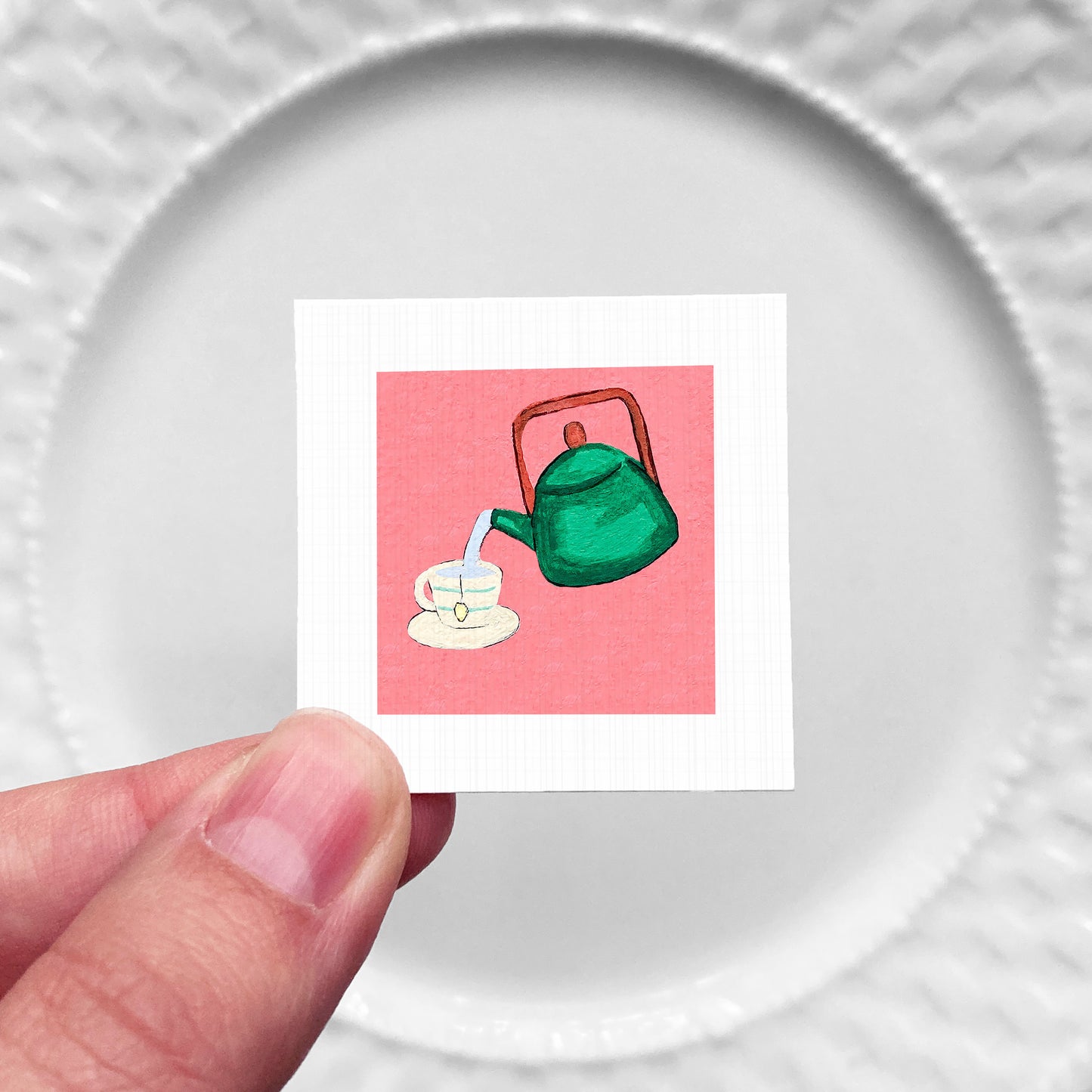 A hand is holding a small square with a green teapot and a cup of tea on it, against a white background.