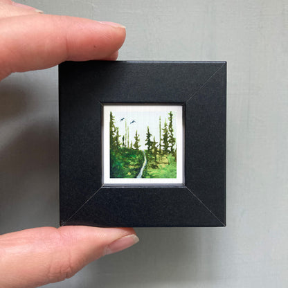A person's hand holding a small black frame containing a painting of a forest scene.