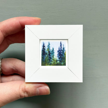 A hand is holding a small white square frame containing a painting of a forest scene with tall trees and a blue sky.