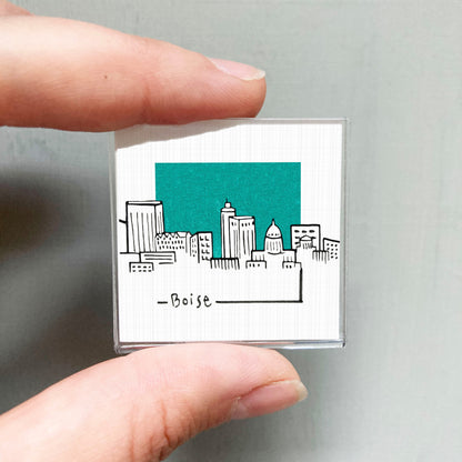 A hand holding a small square-shaped object with a drawing of a city skyline on it.