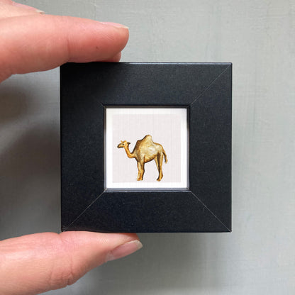 A person's hand is holding a small black frame containing a framed image of a camel.