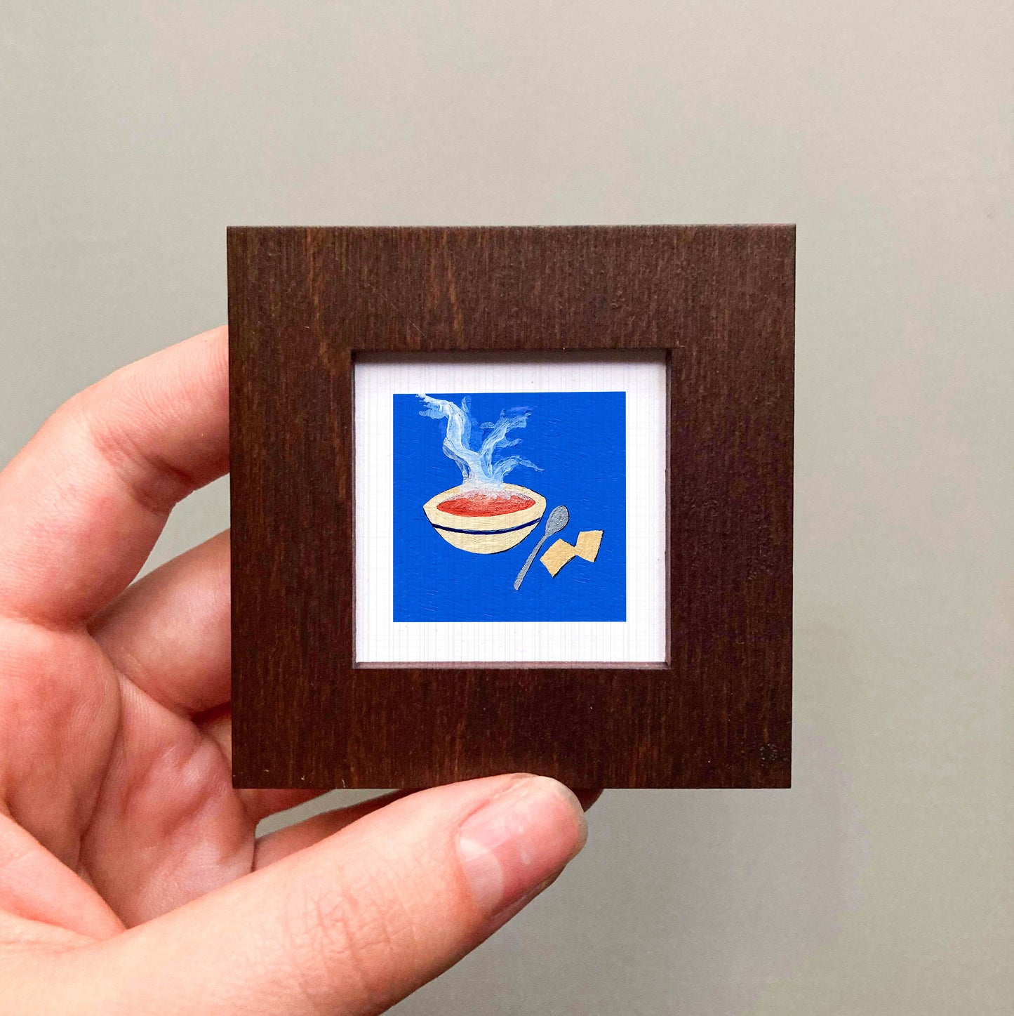 A hand is holding a small square frame containing an illustration of a bowl of soup with steam rising from it.