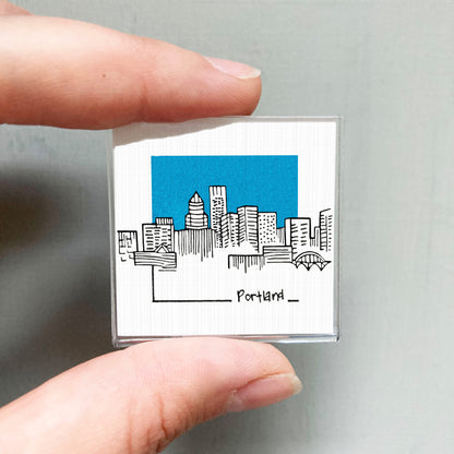 A hand holding a small square magnet with a drawing of a city skyline and the word "Portland" written below it.