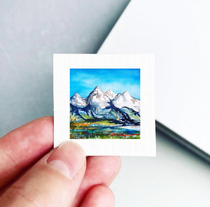 A hand is holding a small square artwork depicting a mountain landscape with snow-capped peaks and a blue sky.