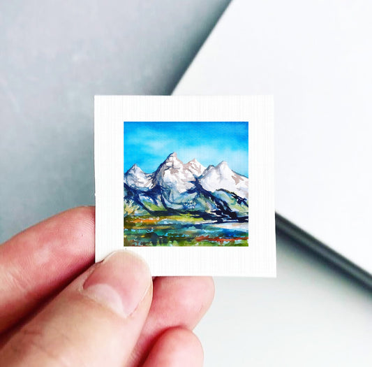 A hand is holding a small square artwork depicting a mountain landscape with snow-capped peaks and a blue sky.