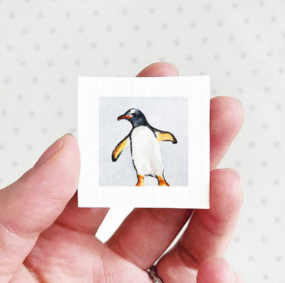A hand holding a small square picture of a penguin against a white background.