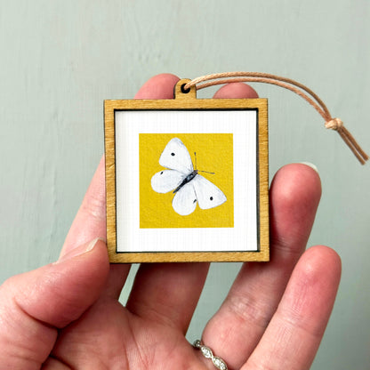 A hand holding a small wooden frame containing a painting of a white butterfly against a yellow background.