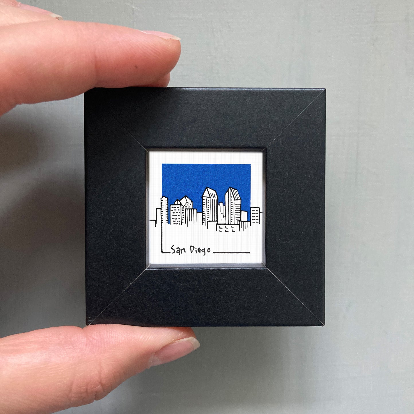 A person's hand holding a small square frame containing an illustration of a city skyline.