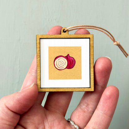 A hand holding a small wooden frame with a picture of a red onion inside it.