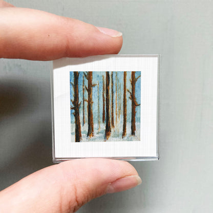 A person's hand holding a small square picture of a snowy forest scene.