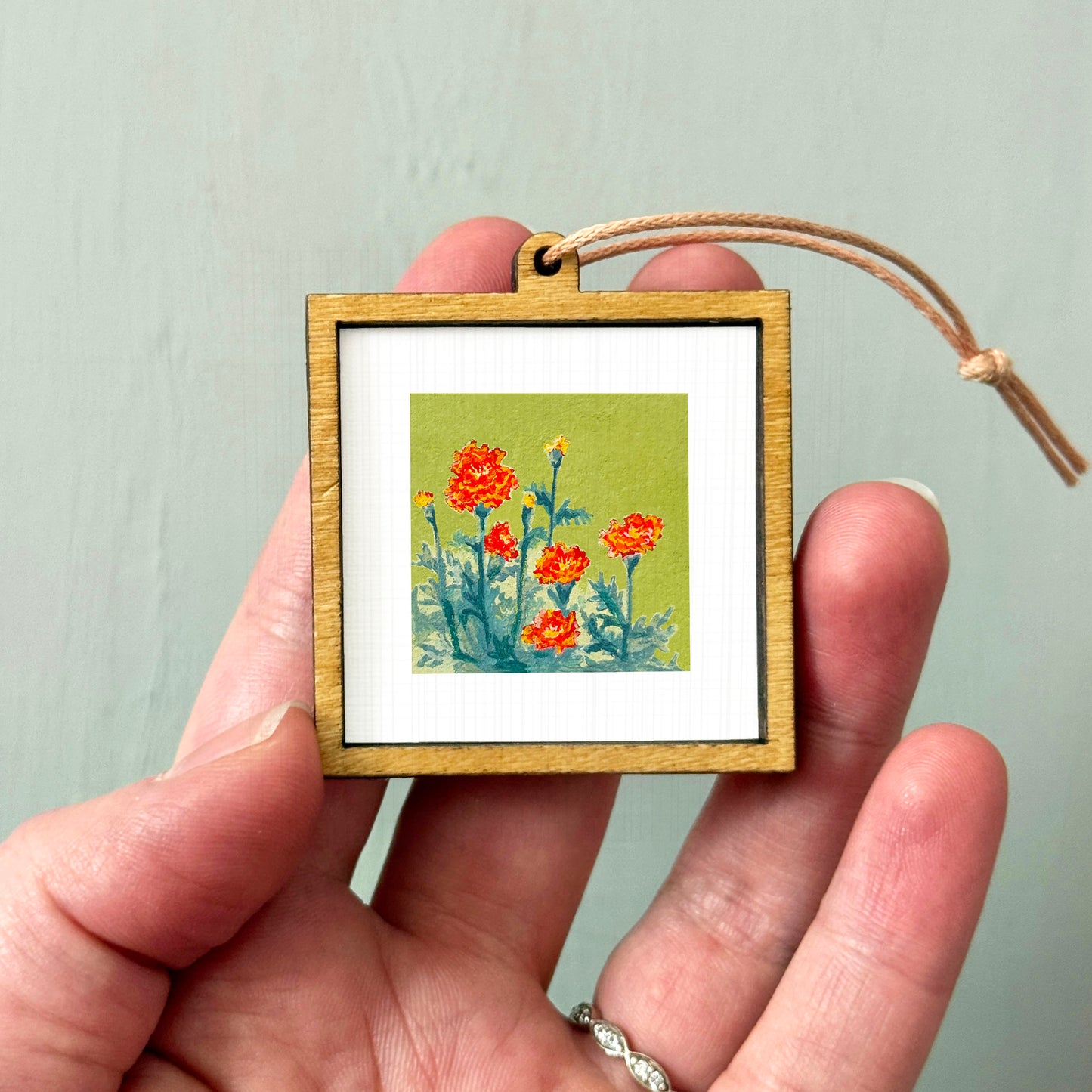A hand holding a small wooden frame containing a painting of orange flowers against a green background.