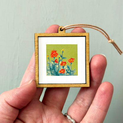 A hand holding a small wooden frame containing a painting of orange flowers against a green background.