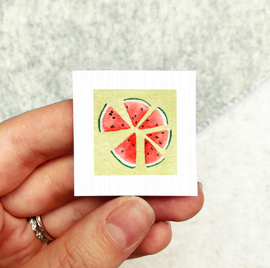A hand holding a small square card with a watermelon slice illustration on it.