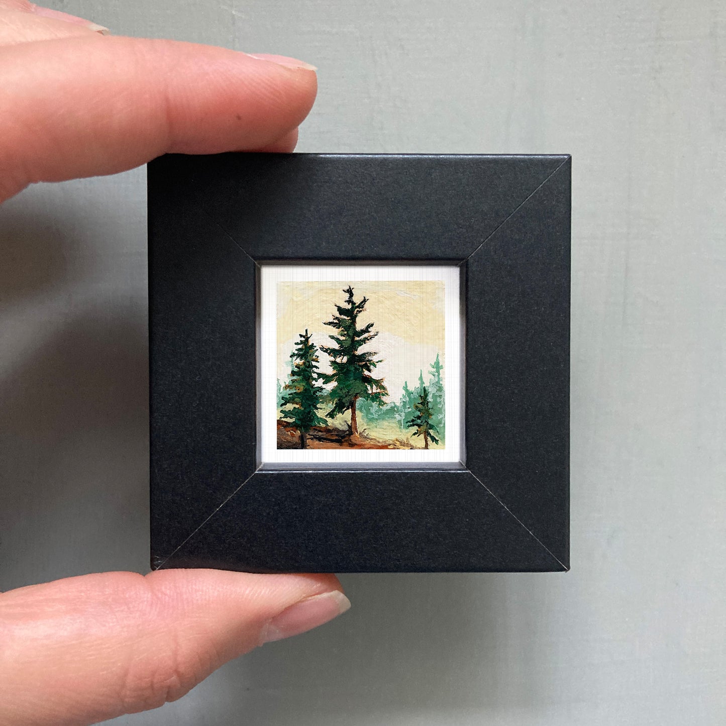 A person's hand holding a small black picture frame containing a painting of a forest scene with pine trees.