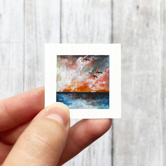 A hand is holding a small square painting of a sunset over a body of water, with birds flying in the sky.