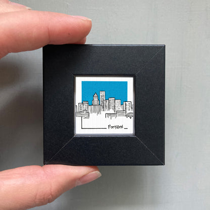 A hand is holding a small black frame containing a framed illustration of a city skyline.