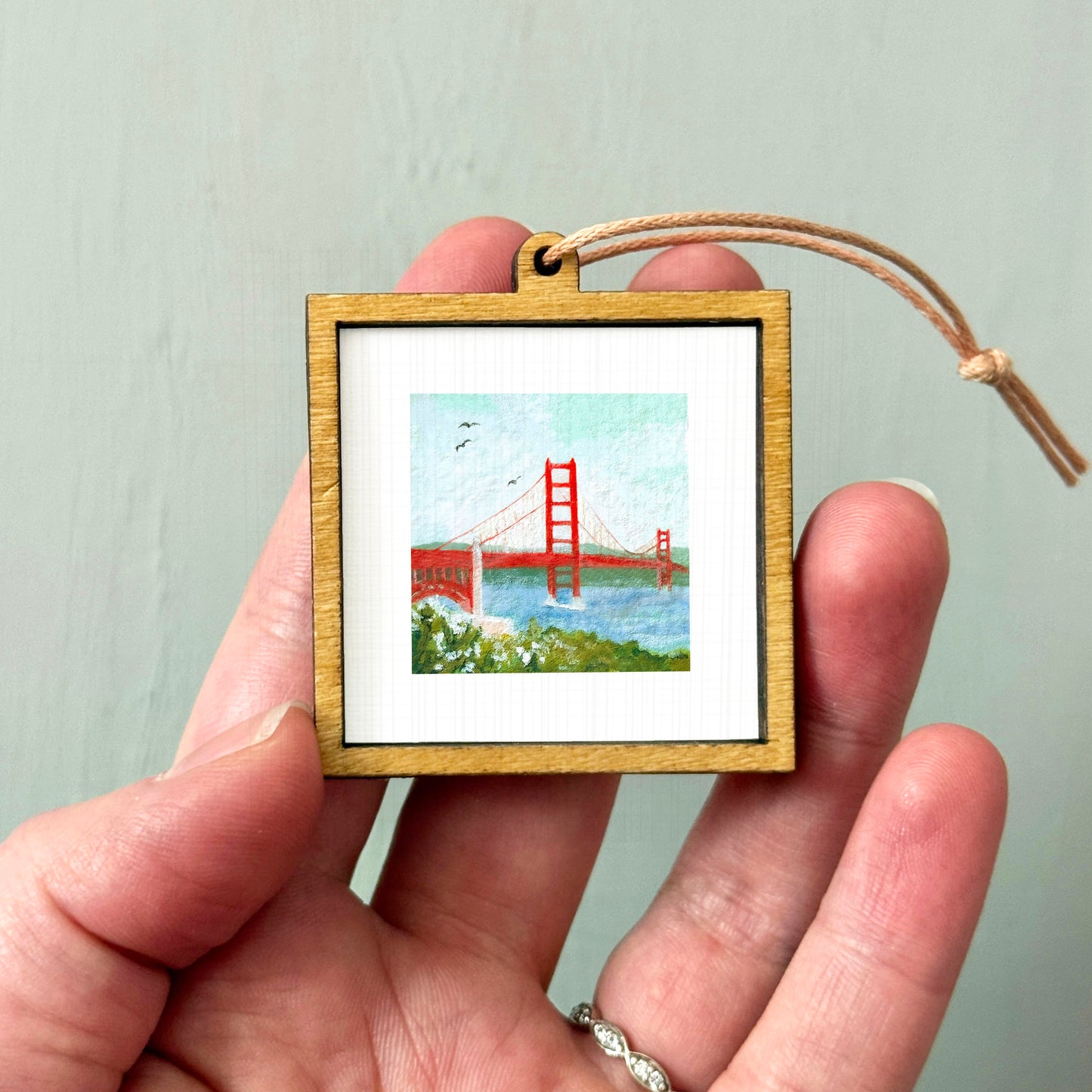 A person's hand holding a small wooden frame containing a painting of the Golden Gate Bridge.