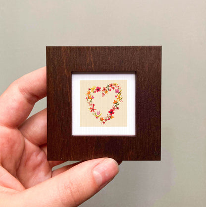 A hand is holding a small wooden frame containing a heart-shaped embroidered design with colorful flowers.