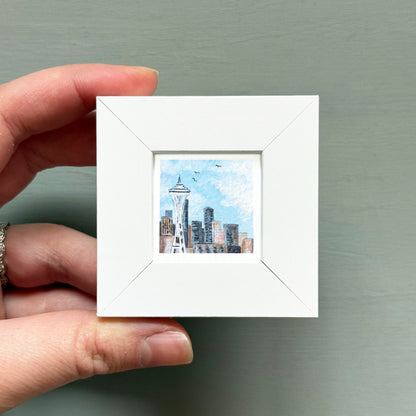 A hand is holding a small white square frame containing a miniature painting of the Seattle skyline.