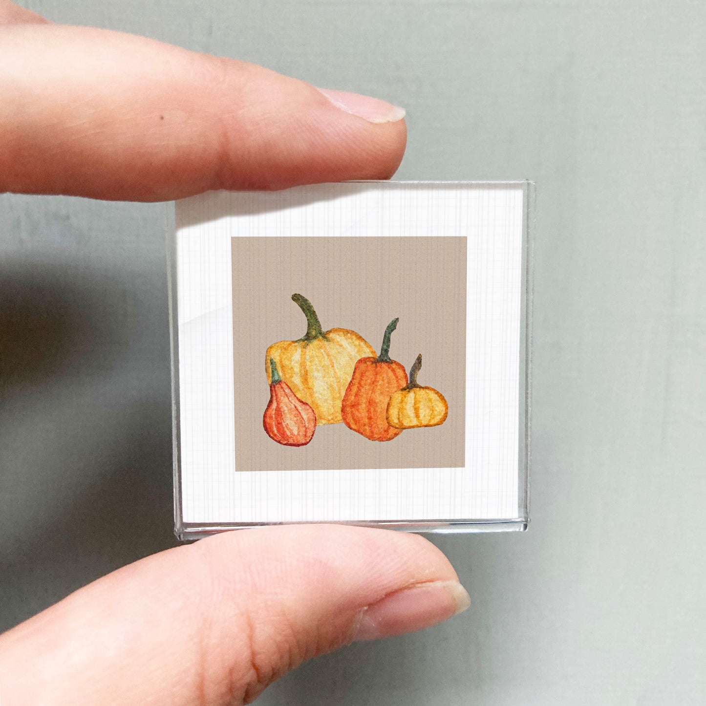 A person's hand holding a small square picture of three pumpkins against a light background.