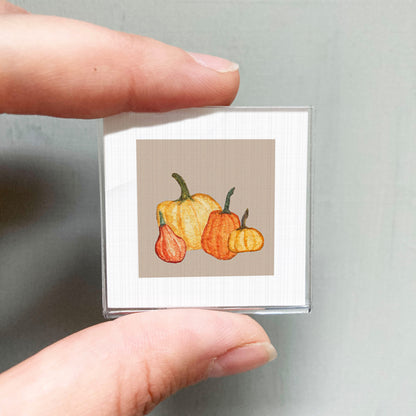 A person's hand holding a small square picture of three pumpkins against a light background.