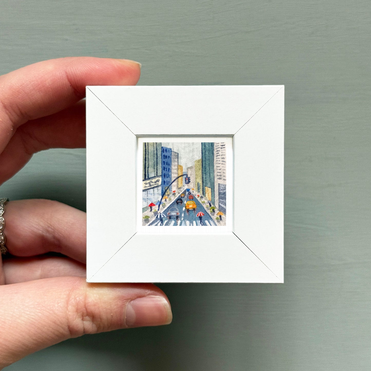 A hand is holding a small white square frame containing a miniature painting of a city street scene.