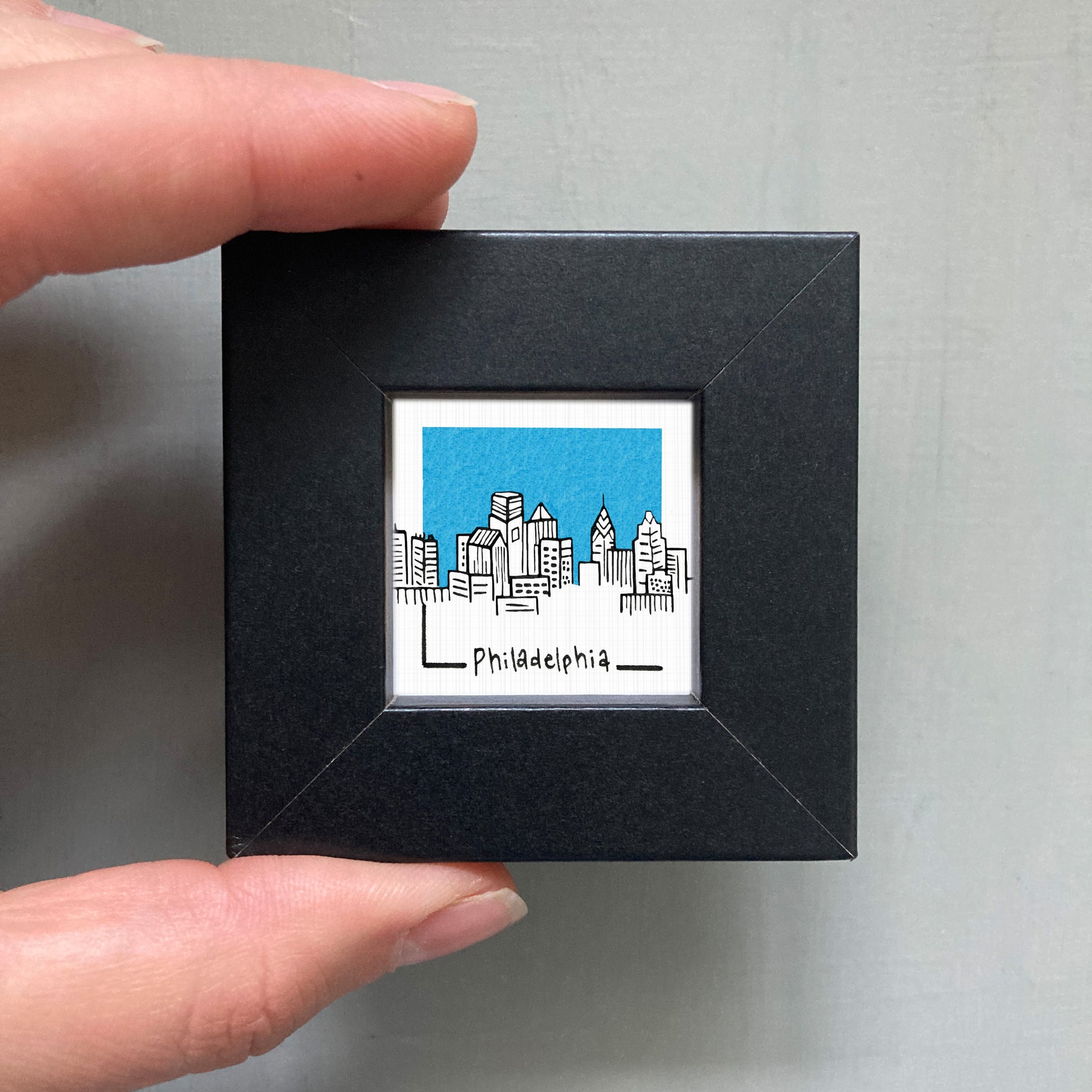 A hand is holding a small black square frame containing a miniature illustration of the Philadelphia skyline.